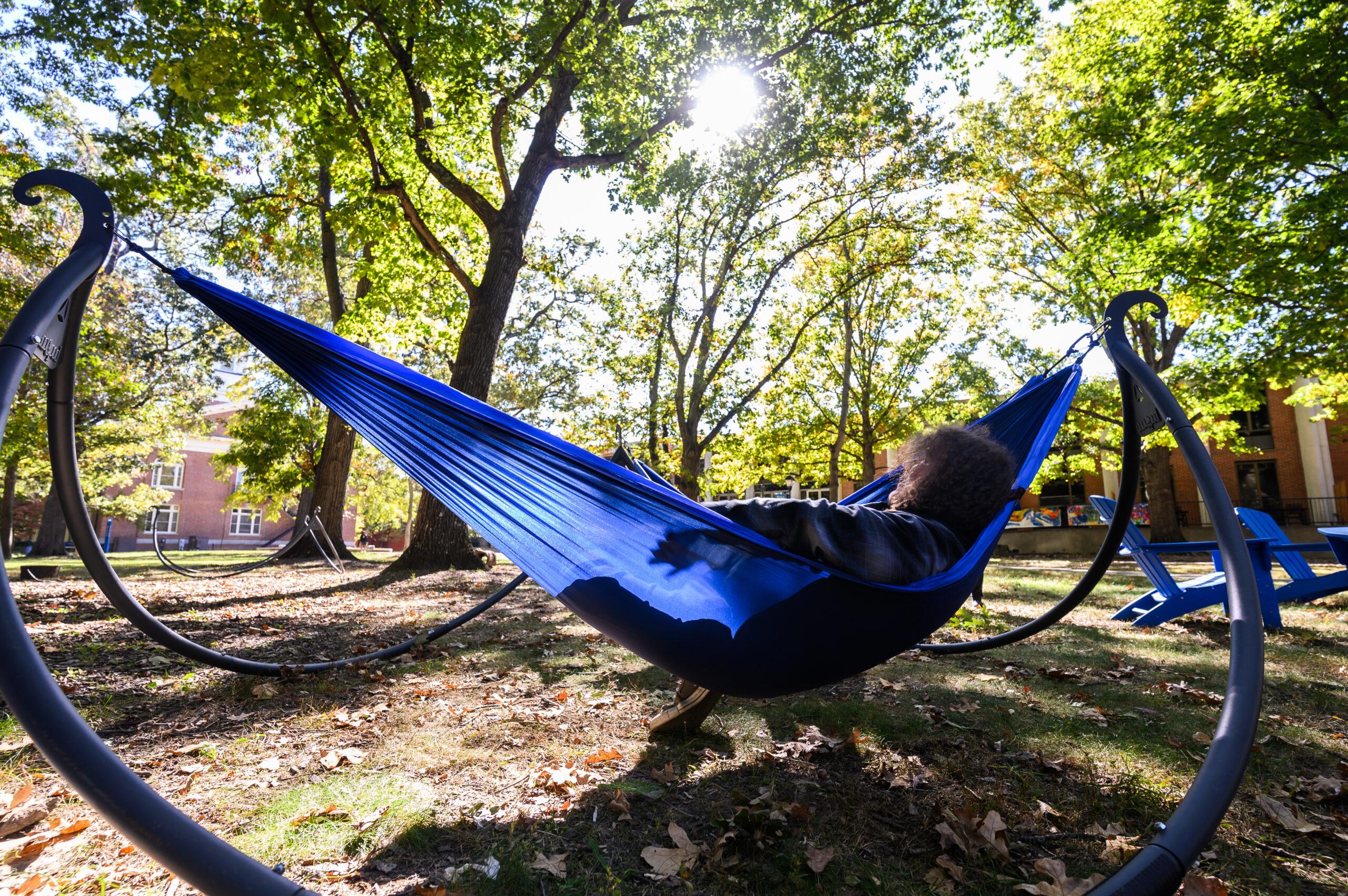 A student is shown lounging in a blue hammock on the quad with the sun casting bright rays of light across the sky above.